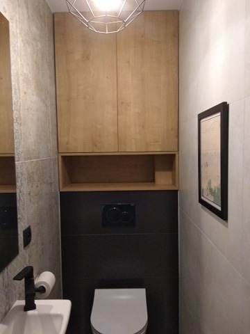 Mała toaleta z drewnianą szafką wiszącą nad zabudową WC i otwartą półką, czarna bateria umywalkowa, geometryczna lampa sufitowa.