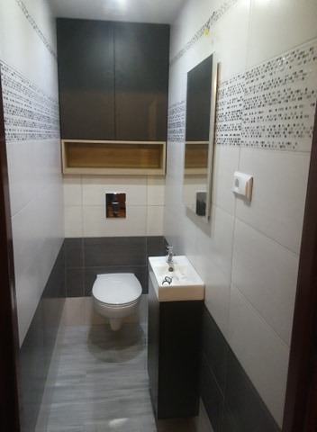 Nowoczesna, wąska łazienka z szarą szafką pod umywalką, wiszącą szafką nad toaletą i lustrem na ścianie z białymi i szarymi płytkami.