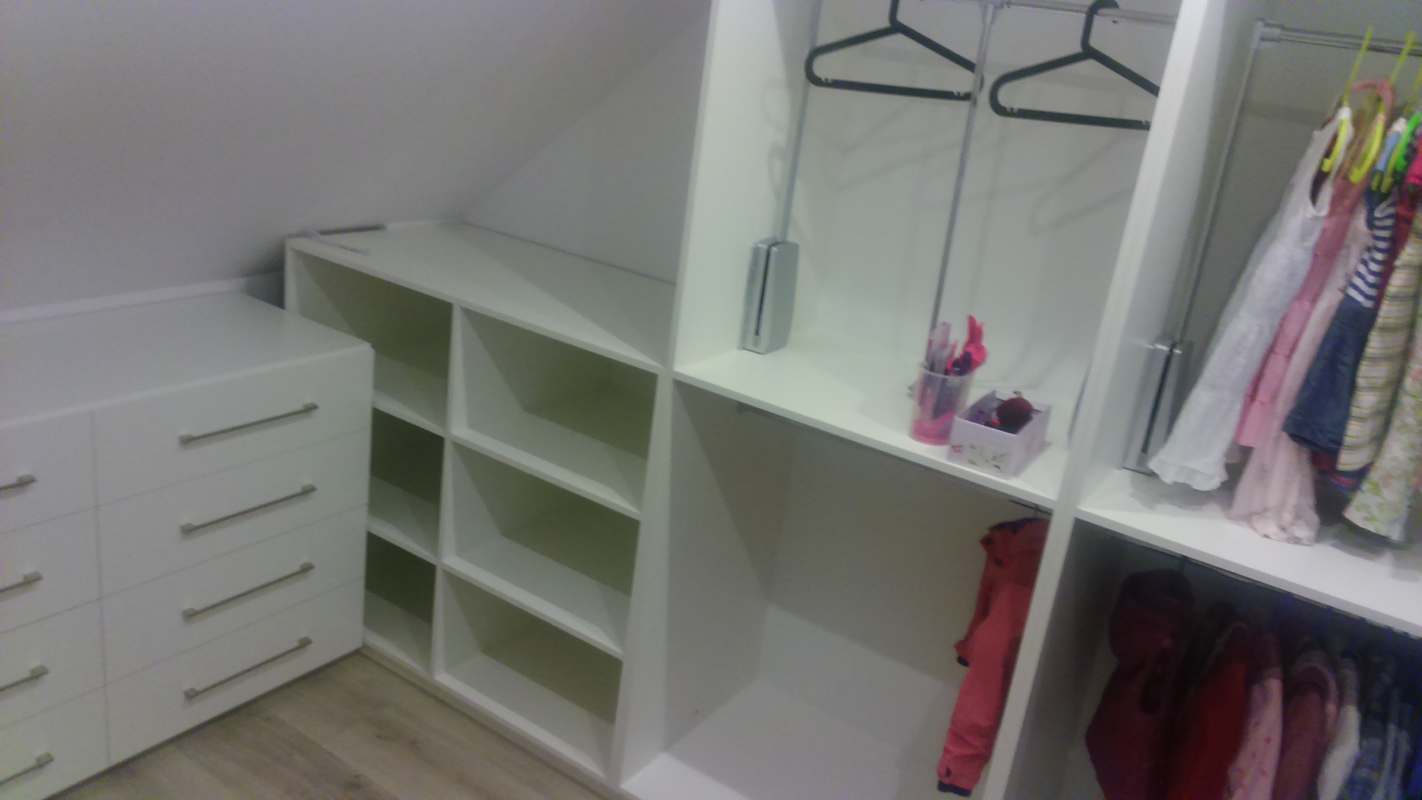 Biała szafa wnękowa pod skosem, z szufladami po lewej stronie i drążkami na ubrania po prawej, częściowo wypełniona ubraniami.