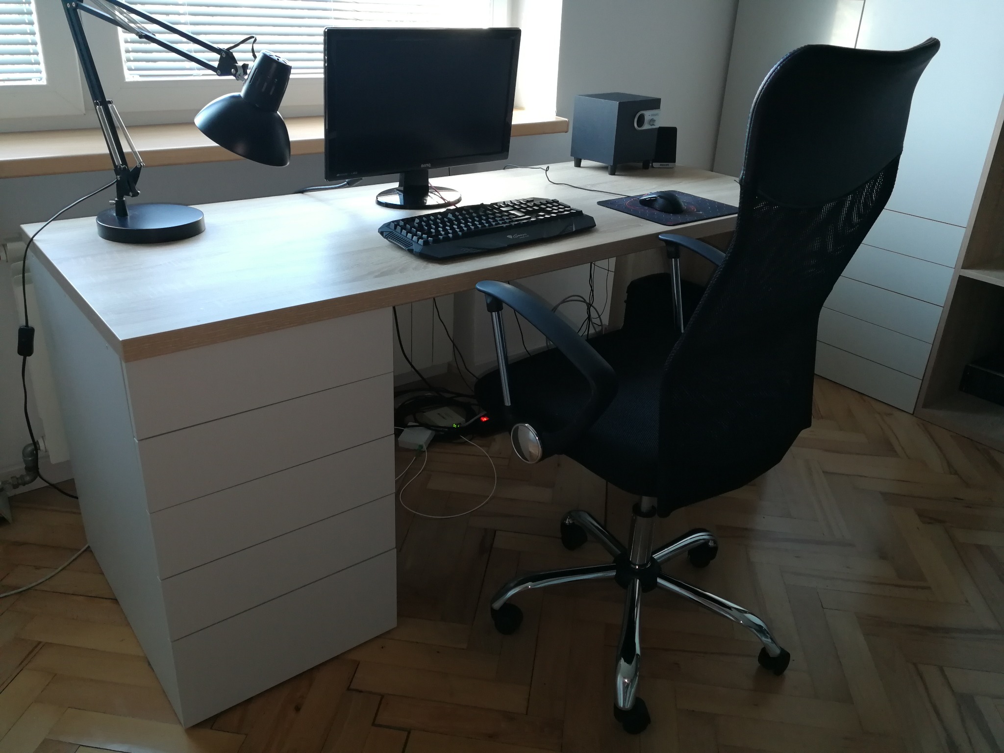 Biurko z blatem drewnianym, szafką z szufladami, lampką, monitorem i krzesłem biurowym na parkiecie.