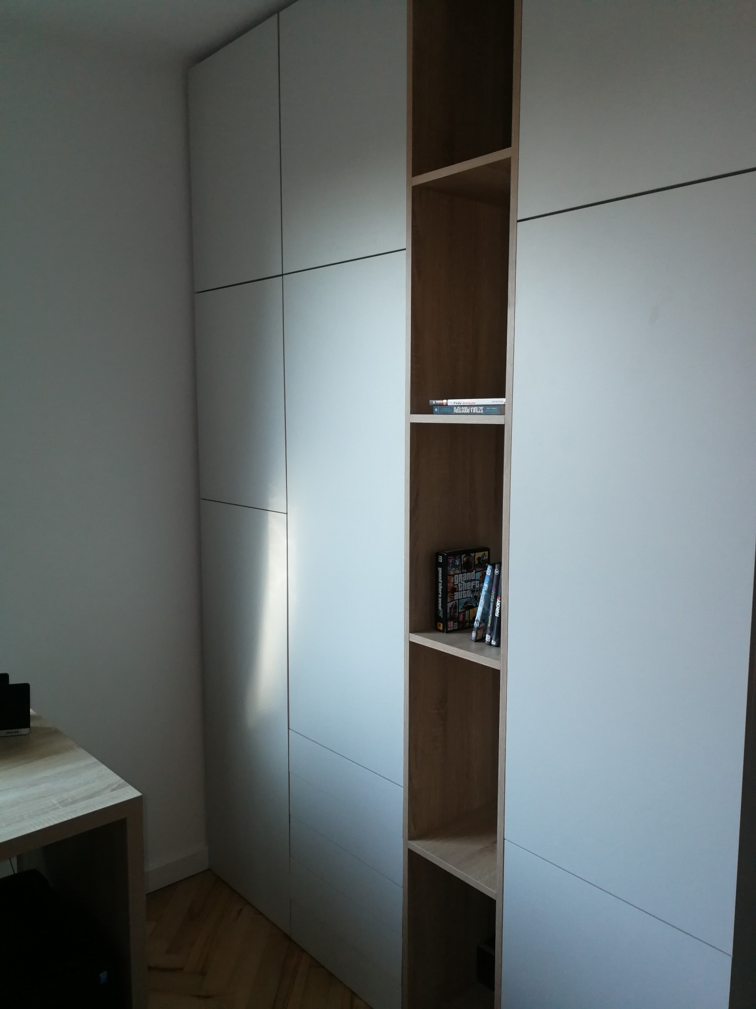 Biała szafa z otwartą półką z książkami i grami, widoczna część biurka z jasnego drewna.