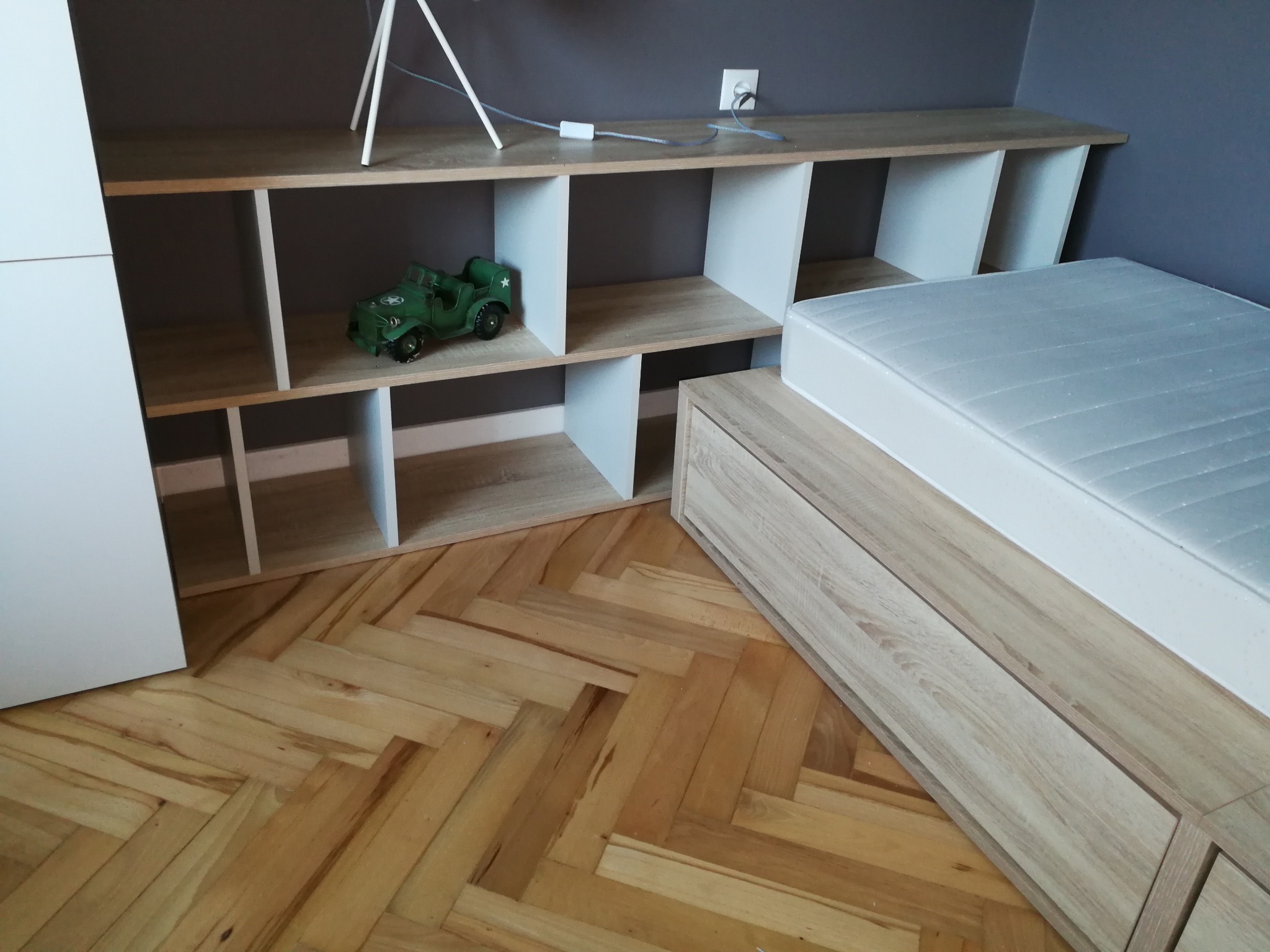 Regał z otwartymi półkami z białymi bokami i drewnianym blatem, ustawiony w pokoju obok łóżka z drewnianą ramą, na podłodze parkiet ułożony w jodełkę, na półce regału stoi zabawkowy zielony jeep.