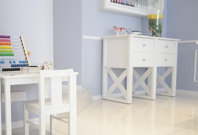 Jasne wnętrze pokoju dziecięcego z białym biurkiem, krzesłem i regałem z szufladami na nóżkach w kształcie litery X, na ścianie półka z lakierami do paznokci.