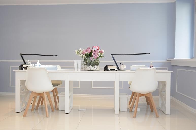 Dwa białe stanowiska do manicure z lampami, narzędziami i kwiatami w wazonie, ustawione w jasnym pomieszczeniu z szarymi ścianami i białymi listwami przypodłogowymi.