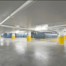 Wnętrze parkingu podziemnego z kolumnami pomalowanymi na żółto, widoczne rury pod sufitem i betonowa posadzka.