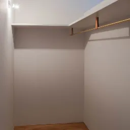 Wnętrze garderoby z drewnianą podłogą, półkami pod sufitem i złotym drążkiem na ubrania, oświetlone punktowo.