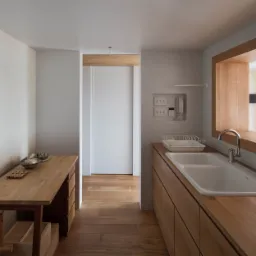 Minimalistyczna kuchnia z drewnianymi szafkami i blatem, białą ścianą, zlewem i widokiem na salon przez okno w ścianie.