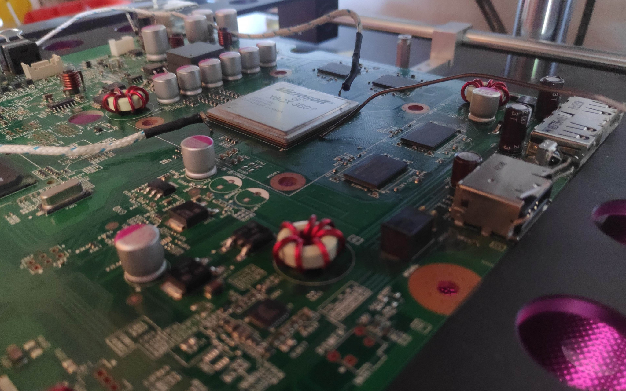 Płyta główna konsoli Xbox 360 z widocznym procesorem Microsoft, elementami elektronicznymi i złączami, na czarnym tle z fioletowym podświetleniem.