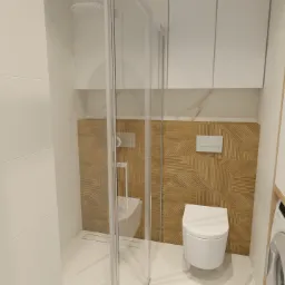 Nowoczesna łazienka z kabiną prysznicową typu walk-in, toaletą podwieszaną i drewnopodobną ścianą dekoracyjną z geometrycznym wzorem, uzupełniona białą zabudową meblową i pralką.