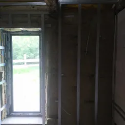 Montaż metalowej konstrukcji pod płyty gipsowo-kartonowe z oknem w tle, widoczna izolacja z wełny mineralnej i drewniany element stropu.