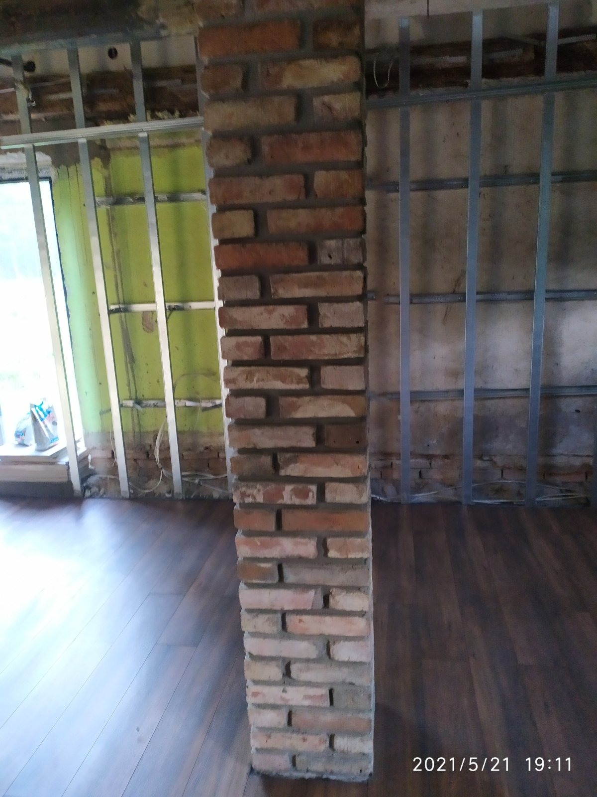 Wnętrze w trakcie remontu: kolumna z cegły klinkierowej, metalowe profile do płyt karton-gips na ścianach, panele podłogowe imitujące drewno.