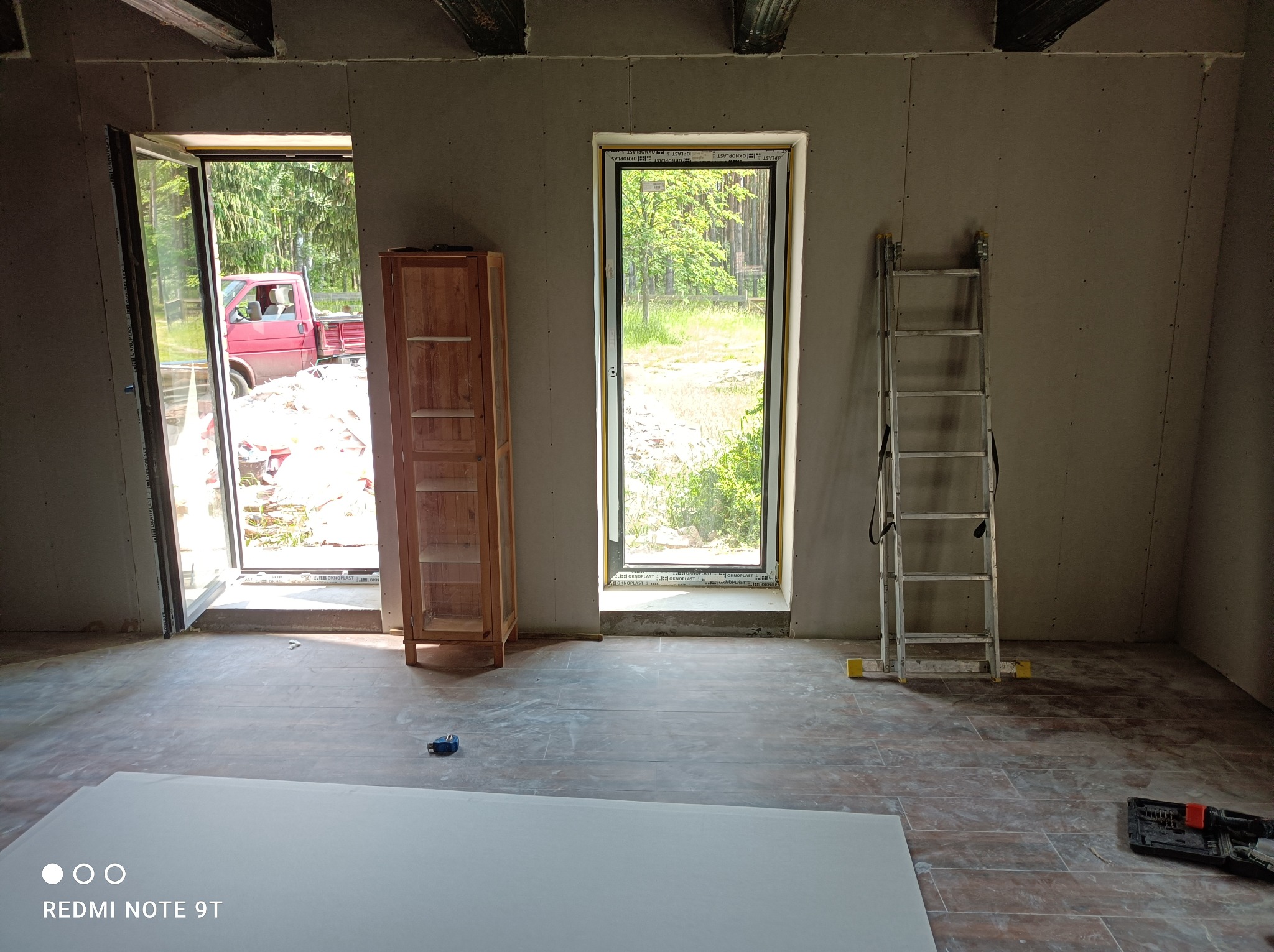 Wnętrze w trakcie prac wykończeniowych: ściany obłożone płytami, widoczne okno i drzwi z widokiem na zewnątrz, drabina oparta o ścianę, fragment szafki, narzędzia i płyta na podłodze.