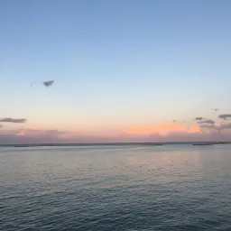 Spokojna powierzchnia wody pod błękitnym i różowym niebem o zachodzie słońca, z delikatnymi falami i odległymi konstrukcjami na horyzoncie.