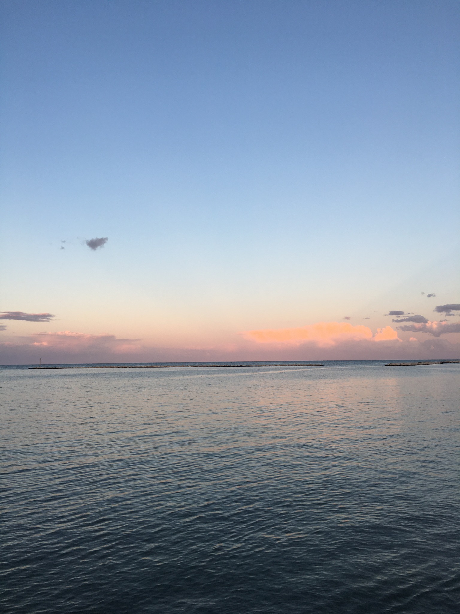 Spokojna powierzchnia wody pod błękitnym i różowym niebem o zachodzie słońca, z delikatnymi falami i odległymi konstrukcjami na horyzoncie.