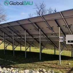 Widok od tyłu na panele słoneczne zamontowane na metalowej konstrukcji, z widocznym inwerterem i puszkami przyłączeniowymi, na tle trawnika i drzew bez liści, z logo firmy w górnym rogu.