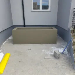 Świeżo otynkowana, prostokątna donica na zewnątrz budynku, z widocznymi śladami zaprawy na podłożu. W tle jasnoszara ściana z oknem i rurą spustową.