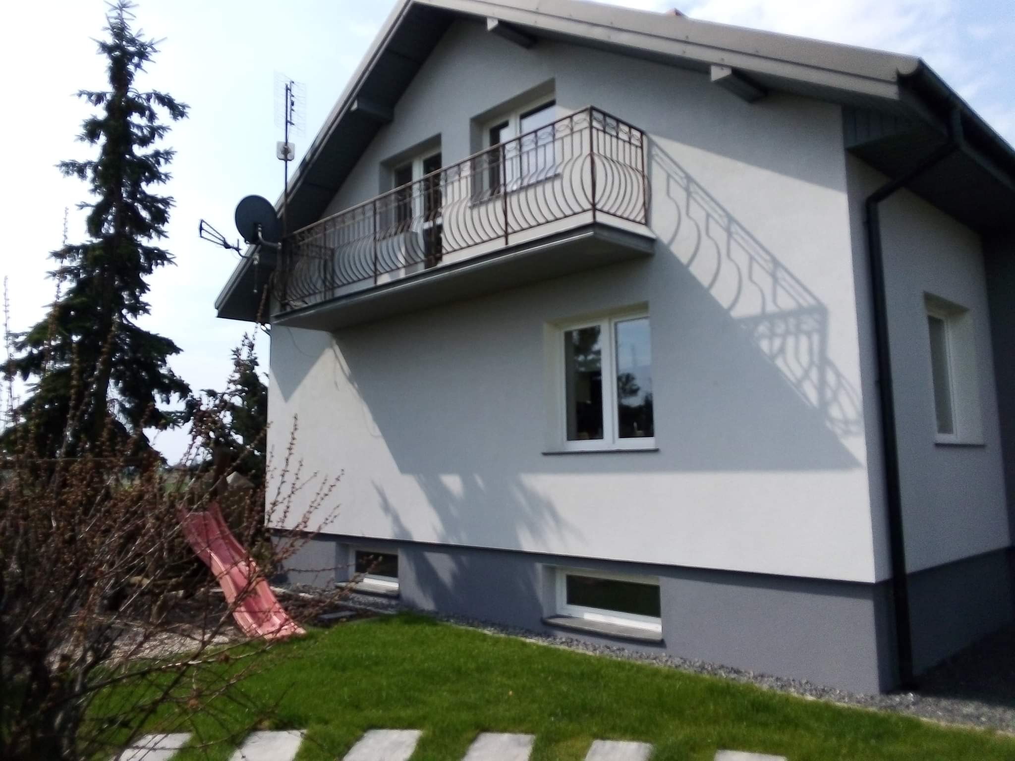 Świeżo otynkowana elewacja domu jednorodzinnego z balkonem i anteną satelitarną, widoczna z poziomu ogrodu z elementami placu zabaw.
