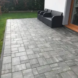 Szara kostka brukowa ułożona na tarasie przy domu, z trzema czarnymi fotelami z tworzywa sztucznego ustawionymi pod ścianą.