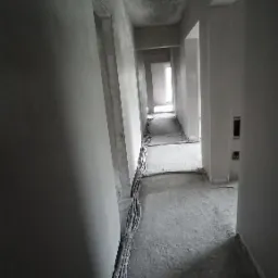 Korytarz w stanie surowym po tynkowaniu maszynowym, widoczne przewody elektryczne przy ścianie, surowy beton na podłodze, światło wpadające z okna na końcu korytarza.