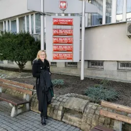 Kobieta w czarnym płaszczu i todze adwokackiej stoi przed budynkiem Sądu Okręgowego w Krakowie, uśmiechając się do kamery.