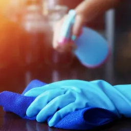 Ręka w niebieskiej rękawicy lateksowej czyści ciemną, błyszczącą powierzchnię niebieską ściereczką z mikrofibry; w tle rozpylacz z płynem do czyszczenia.