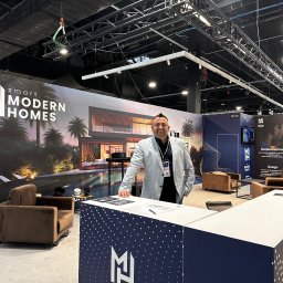 Smart Modern Homes Sp. z o.o. - Stoisko firmy Smart Modern Homes na targach, z uśmiechniętym mężczyzną w jasnej marynarce opartym o ladę z logo firmy, w tle wizualizacja nowoczesnego domu z basenem i palmami.