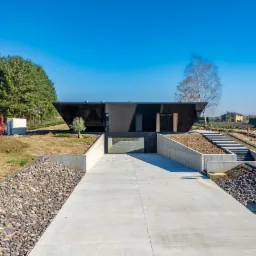 Nowoczesny, czarny dom z garażem podziemnym i betonowym podjazdem, otoczony przygotowanym terenem pod ogród.