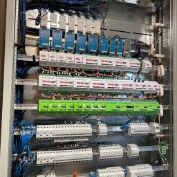 Smart Modern Homes Sp. z o.o. - Rozdzielnica elektryczna z modułami sterującymi, widoczne liczne przewody, moduły zasilające MW NDR-240-24, moduły Nexwell oraz zabezpieczenia Eaton, całość zamontowana w metalowej obudowie.