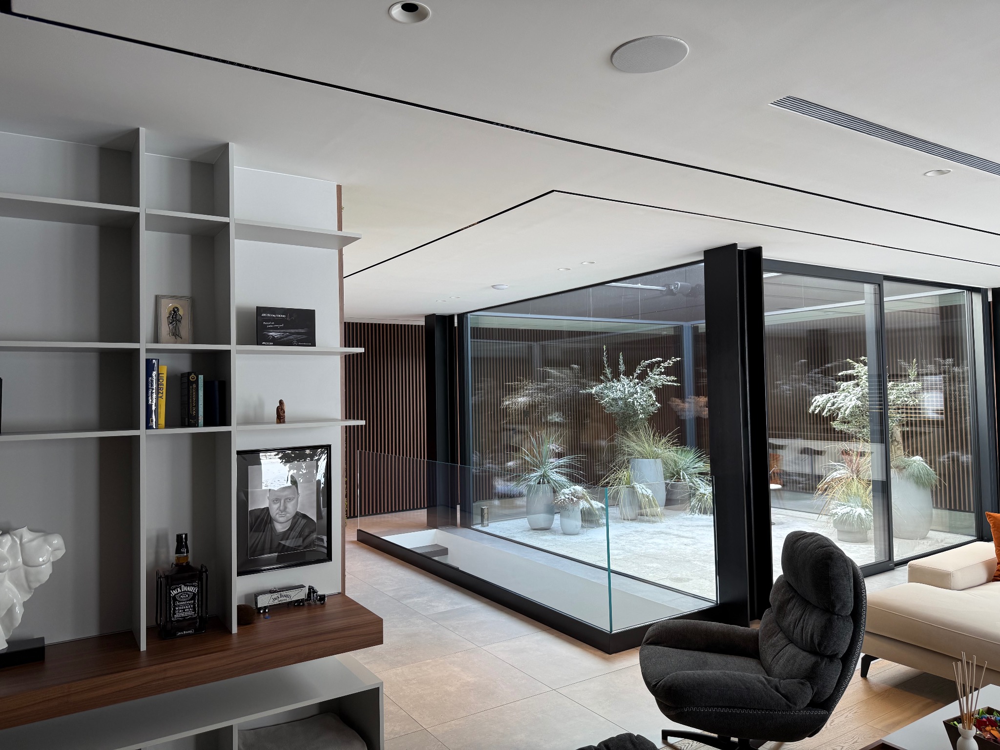 Nowoczesne wnętrze salonu z przeszklonym patio, widoczne rośliny w donicach, minimalistyczny design, szare meble, półki z dekoracjami i alkoholem Jack Daniels.