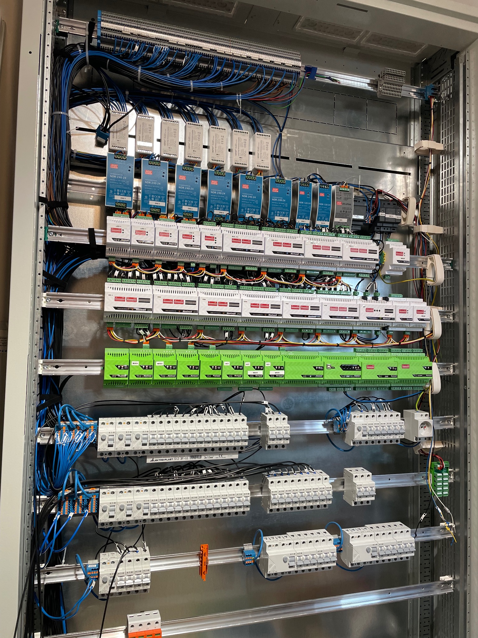 Rozdzielnica elektryczna z modułami sterującymi, widoczne liczne przewody, moduły zasilające MW NDR-240-24, moduły Nexwell oraz zabezpieczenia Eaton, całość zamontowana w metalowej obudowie.
