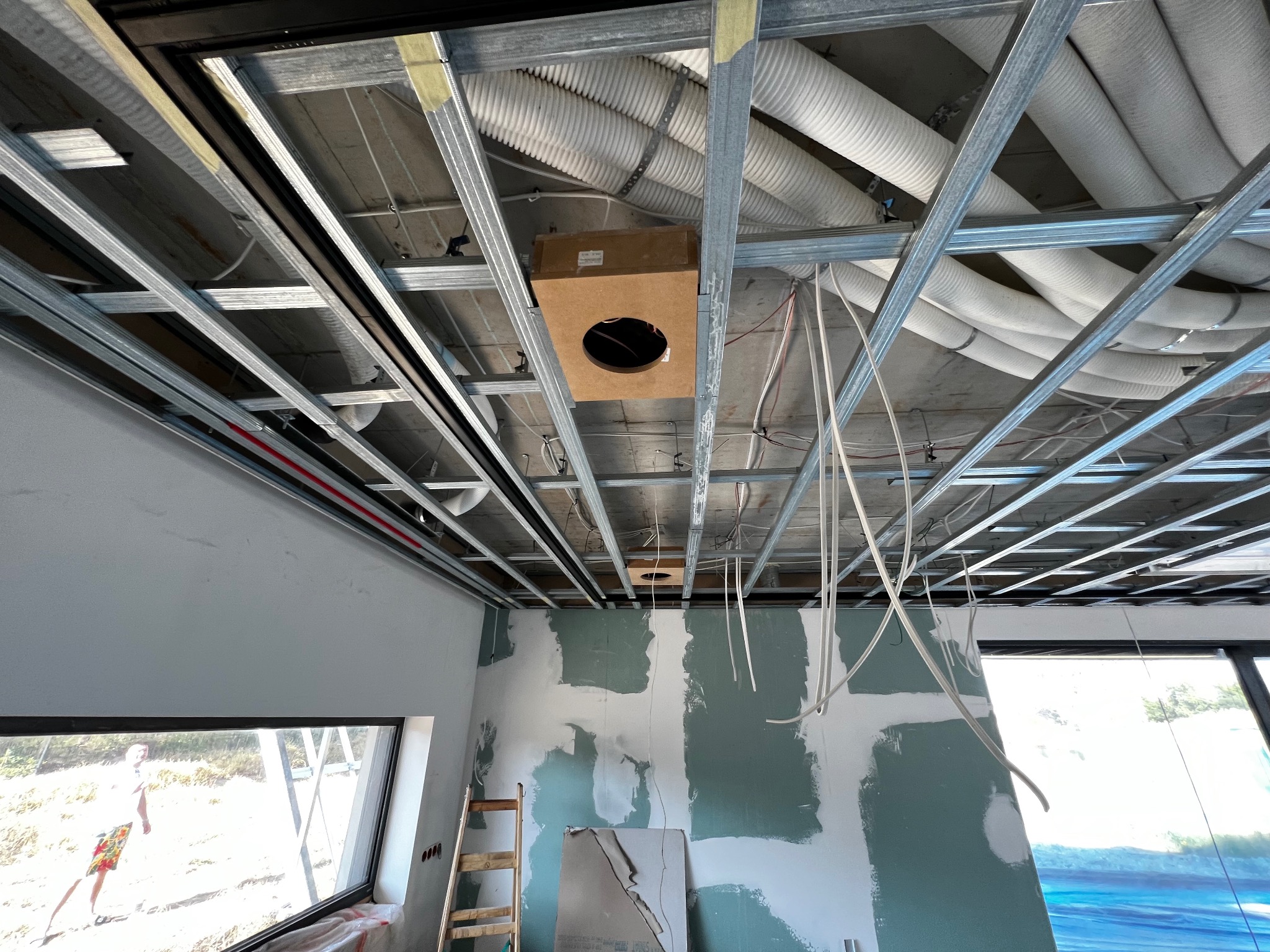 Instalacja podwieszanego sufitu z widocznymi rurami wentylacyjnymi, przewodami elektrycznymi oraz głośnikiem w drewnianej obudowie, ściany w trakcie szpachlowania.