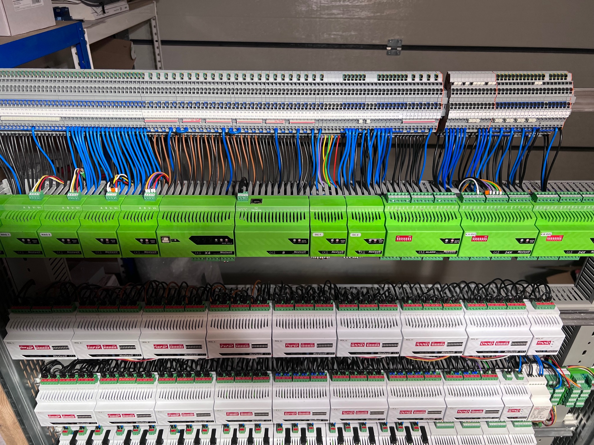 Panel sterowania automatyką budynku z zielonymi modułami Nexwell i licznymi przewodami w kolorach niebieskim, czarnym i brązowym, podłączonymi do listew zaciskowych