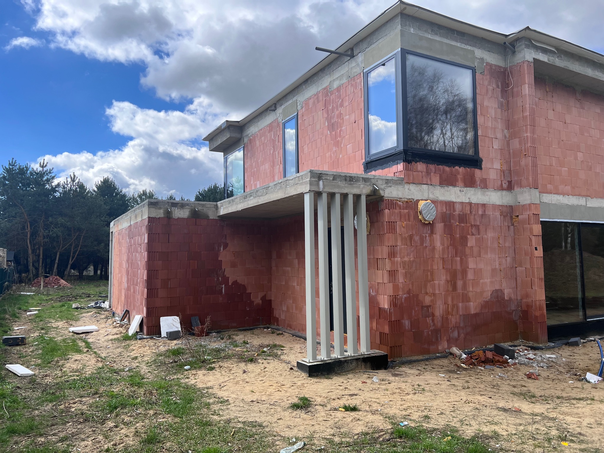 Surowy stan domu jednorodzinnego z czerwonej cegły ceramicznej, widoczne duże okna z czarnymi ramami, betonowe elementy konstrukcyjne, niezabezpieczona działka z piaskiem i resztkami materiałów...