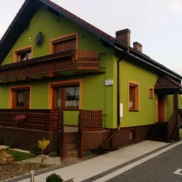 Zakończona realizacja: Dom jednorodzinny z zieloną elewacją, brązowymi detalami (okna, balustrady) i numerem 76, widok z zewnątrz na posesję z kostki brukowej.