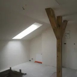 Wnętrze poddasza w trakcie prac wykończeniowych, widoczna drewniana konstrukcja wspierająca, okno dachowe, instalacja elektryczna oraz przygotowane podłoże pod wylewkę.