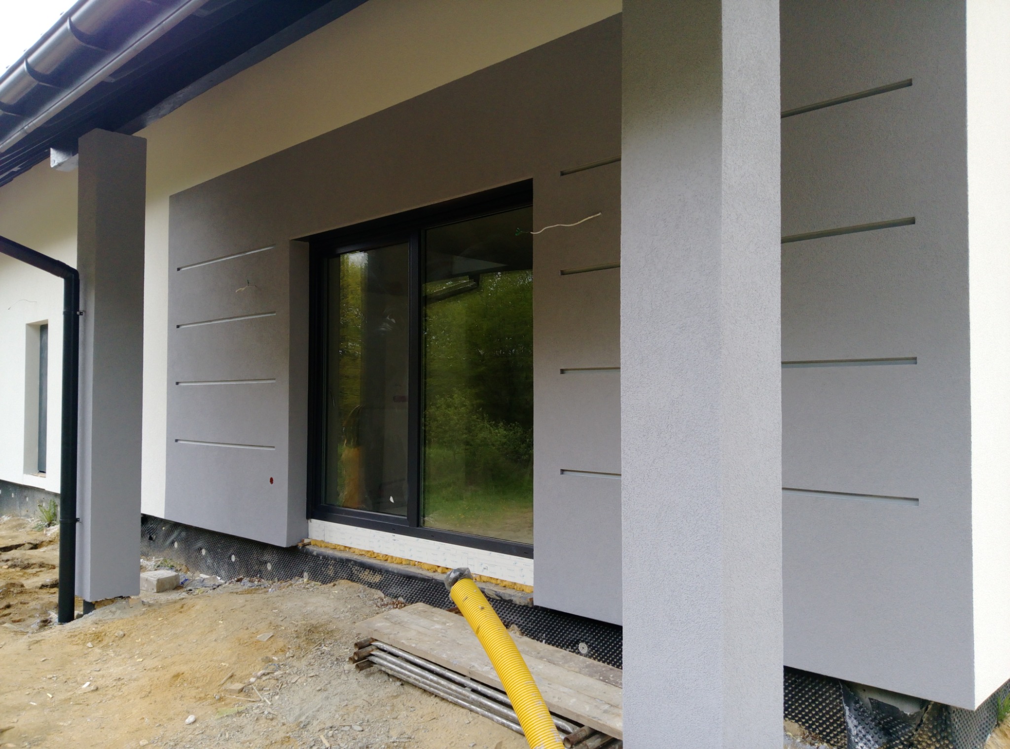 Elewacja domu jednorodzinnego w trakcie prac wykończeniowych, widoczna żółta rura wentylacyjna i fragment terenu budowy.