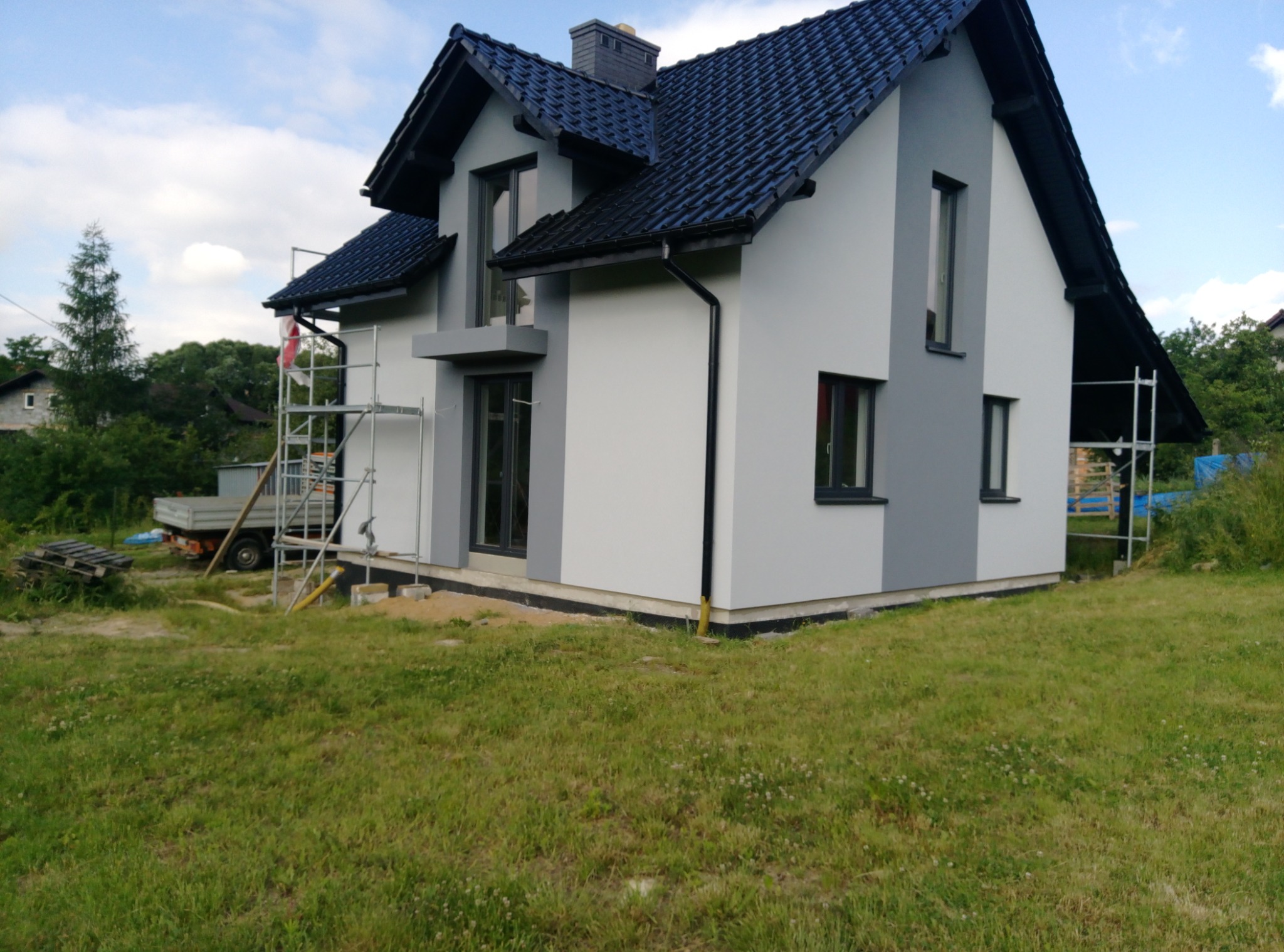 Dwukondygnacyjny dom z szarą elewacją w trakcie prac wykończeniowych, widoczne rusztowania, przygotowany teren wokół budynku.