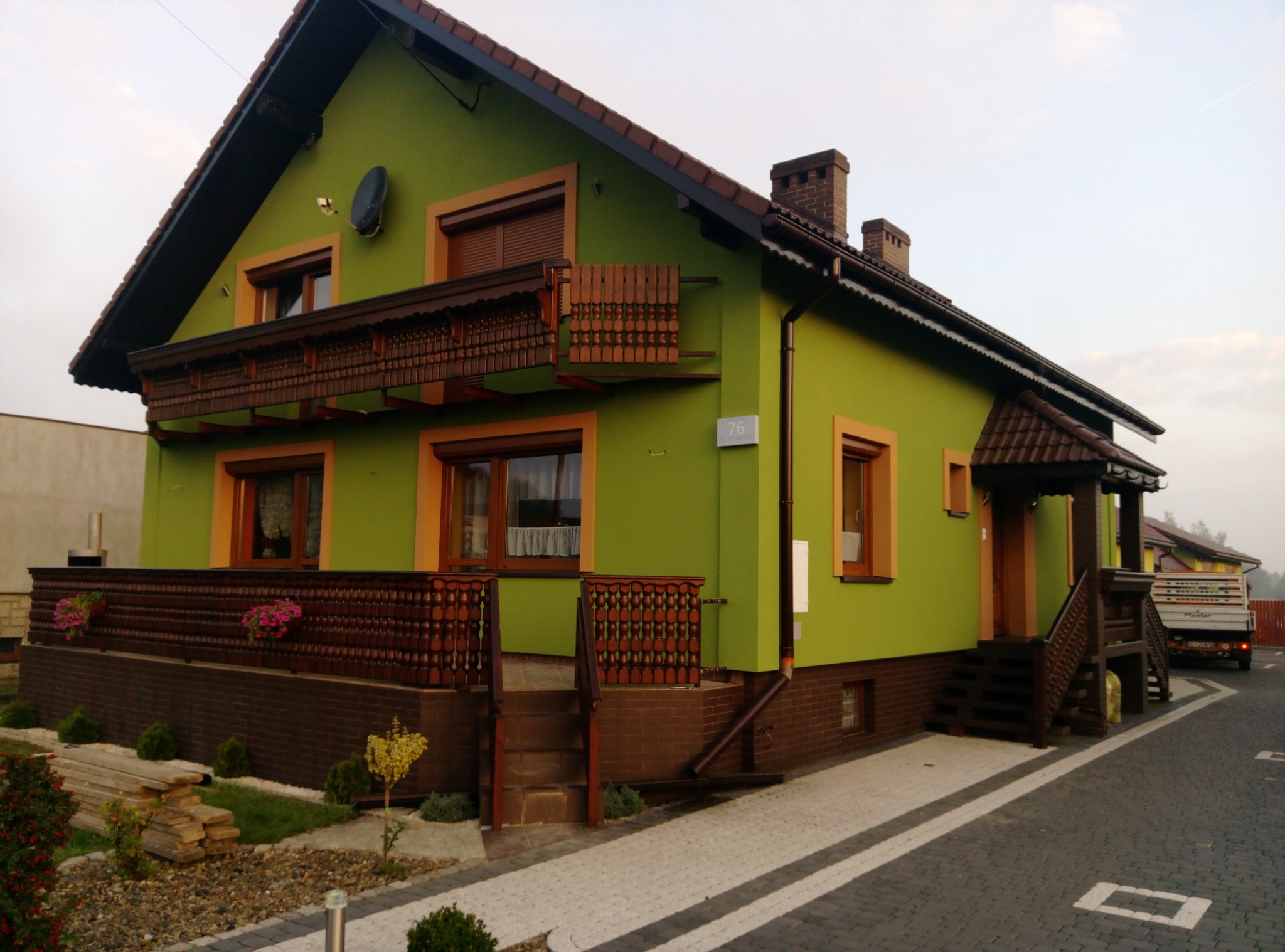 Zakończona realizacja: Dom jednorodzinny z zieloną elewacją, brązowymi detalami (okna, balustrady) i numerem 76, widok z zewnątrz na posesję z kostki brukowej.