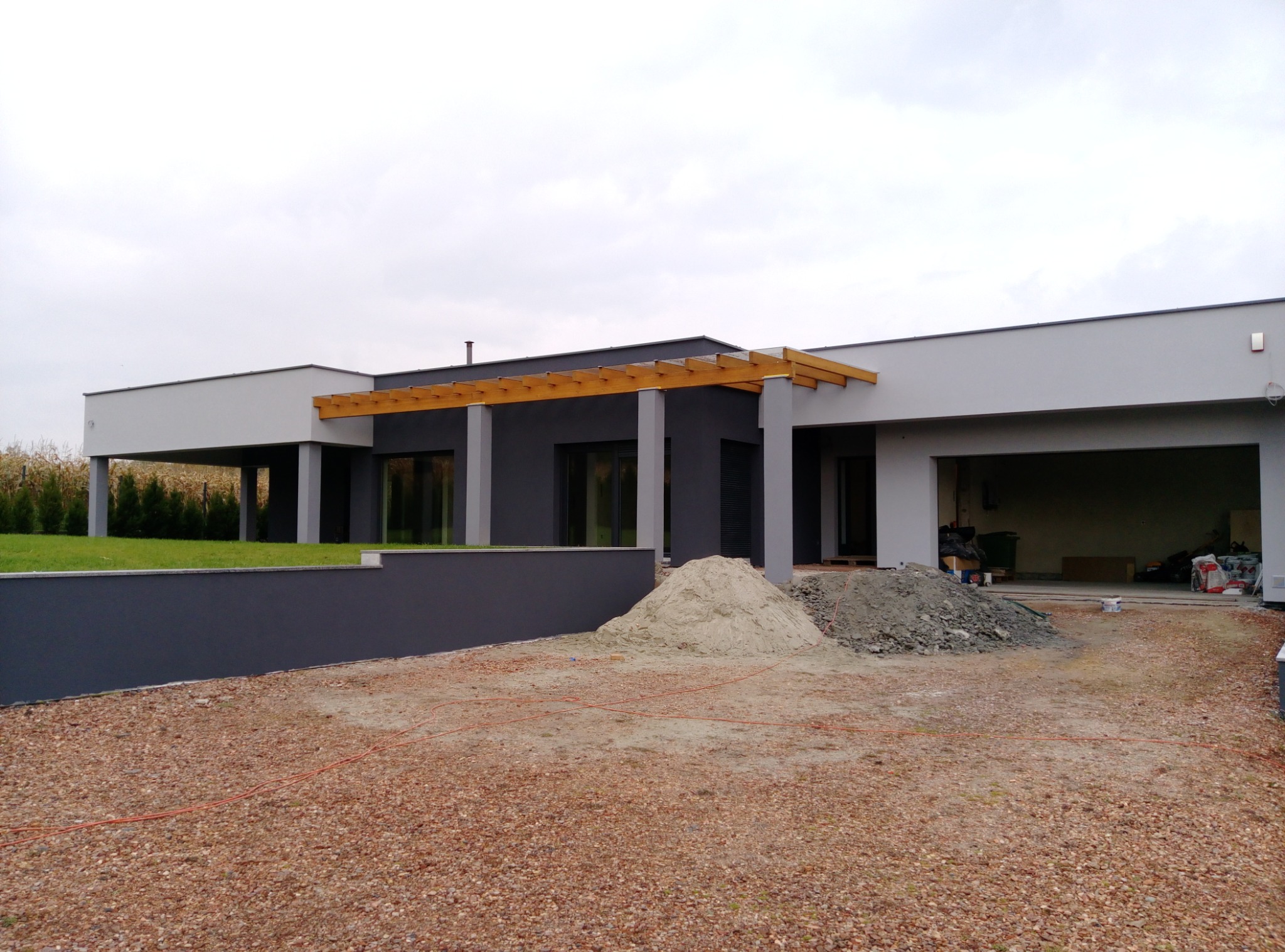 Nowoczesny dom jednorodzinny z szarą elewacją i drewnianą pergolą, widoczne sterty piasku i gruzu przed budynkiem, sugerujące prace wykończeniowe.