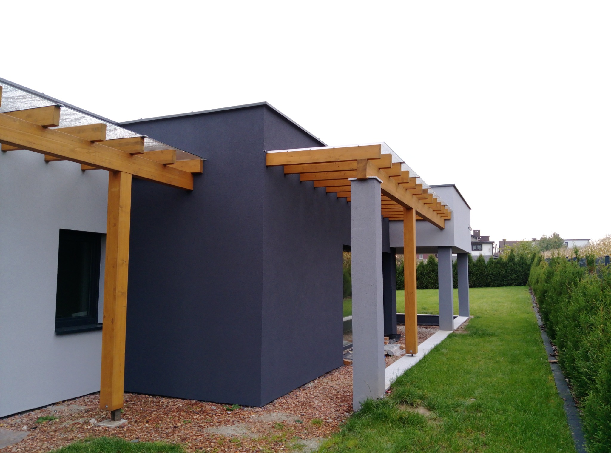 Nowoczesny dom z ciemnoszarą elewacją i drewnianą pergolą z przezroczystym zadaszeniem, widok z boku na trawnik i żywopłot.