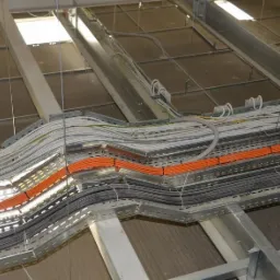 Instalacja elektryczna pod sufitem: koryta kablowe z ułożonymi przewodami w kolorach pomarańczowym, białym i szarym.