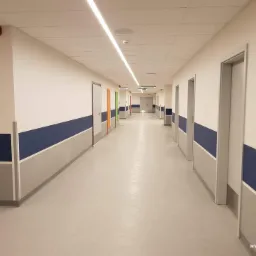 Długi korytarz w budynku użyteczności publicznej z szarą podłogą, białymi ścianami z niebieskim pasem i rzędem drzwi po obu stronach, oświetlony liniowymi lampami sufitowymi.