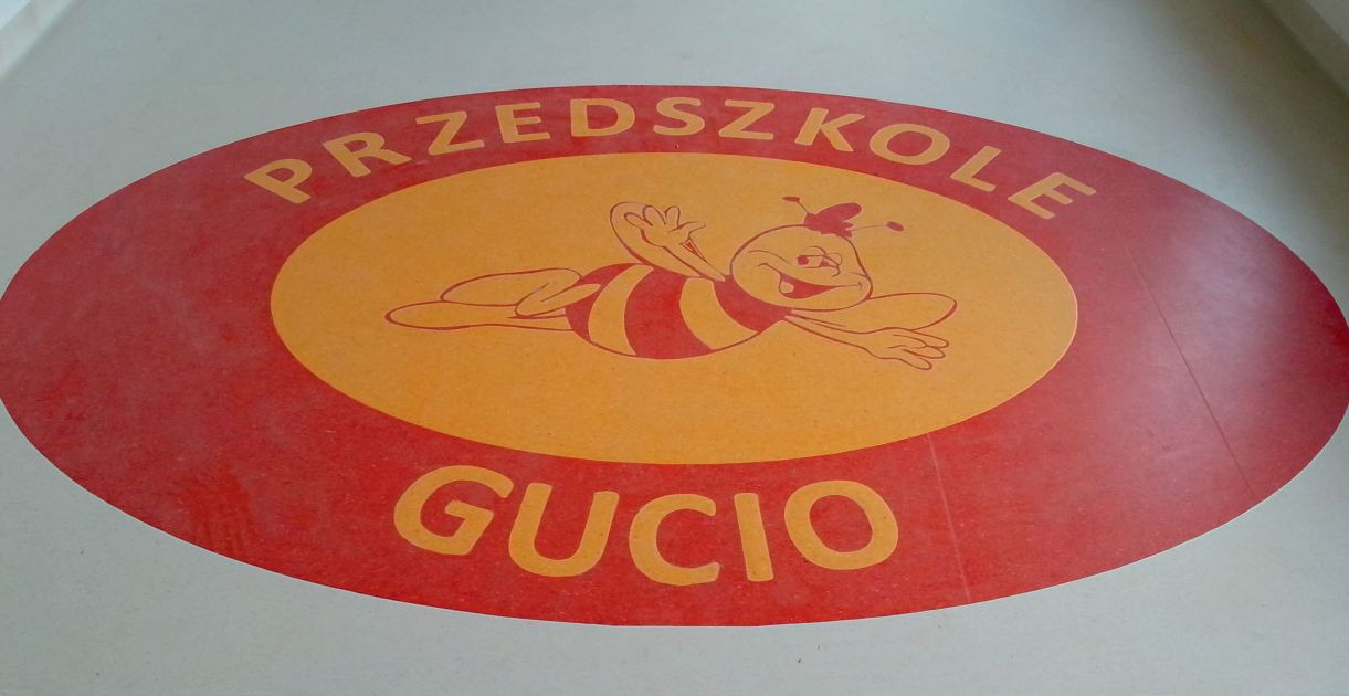 Podłoga w przedszkolu z okrągłym logo przedstawiającym pszczołę Maję i napis 'Przedszkole Gucio' w kolorach czerwonym i pomarańczowym.
