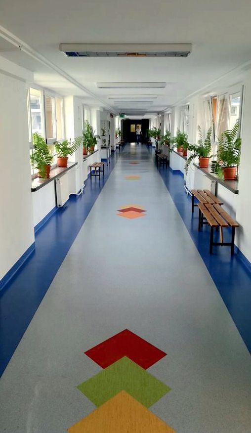 Długi korytarz w budynku użyteczności publicznej z jasnoszarą podłogą z kolorowymi geometrycznymi wzorami, niebieskimi przypodłogowymi listwami, drewnianymi ławkami pod oknami i doniczkowymi...