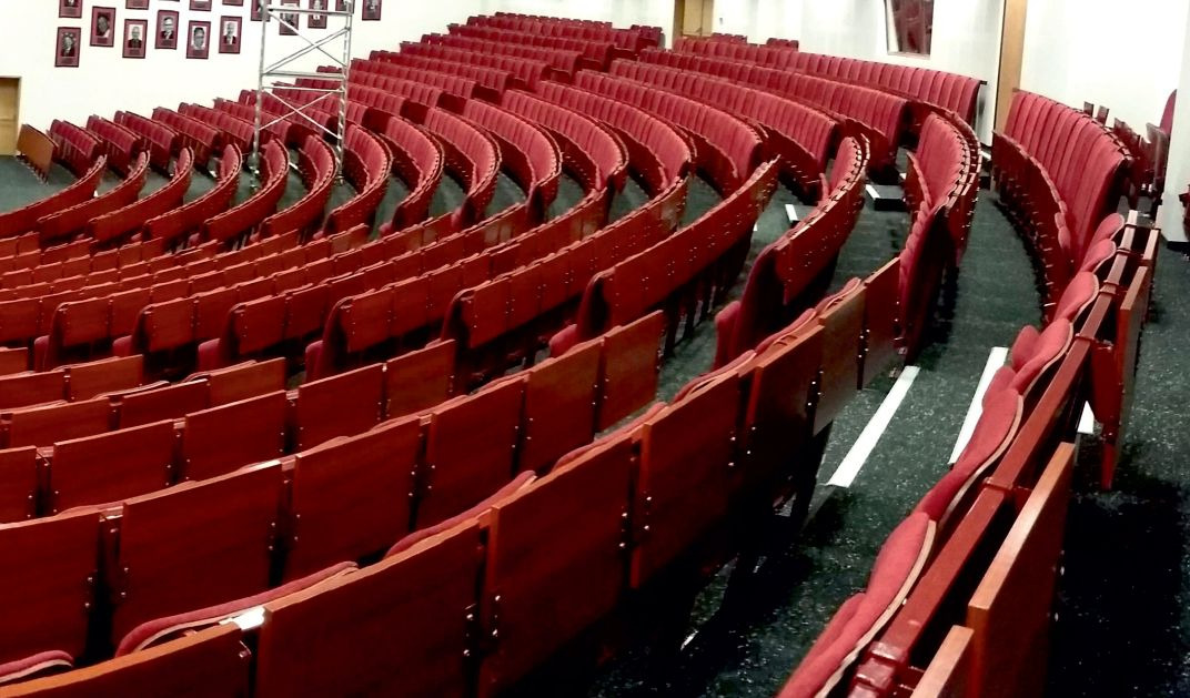 Rzędy czerwonych, składanych krzeseł w audytorium z czarną podłogą i białymi pasami, widoczne rusztowanie i portrety na ścianie w tle.