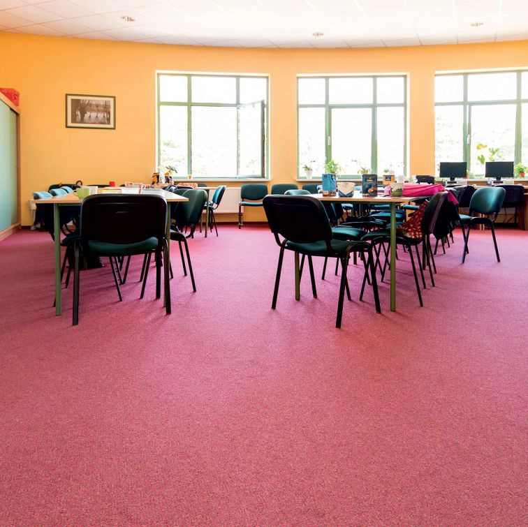Jasnoróżowa wykładzina dywanowa w szkolnej sali z żółtymi ścianami, stolikami i krzesłami, widok na okna i wyposażenie.