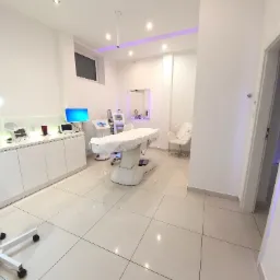 Jasne, przestronne wnętrze gabinetu z białymi meblami, sprzętem kosmetycznym i fotelem, oświetlone lampami sufitowymi i lampą podłogową z lupą.