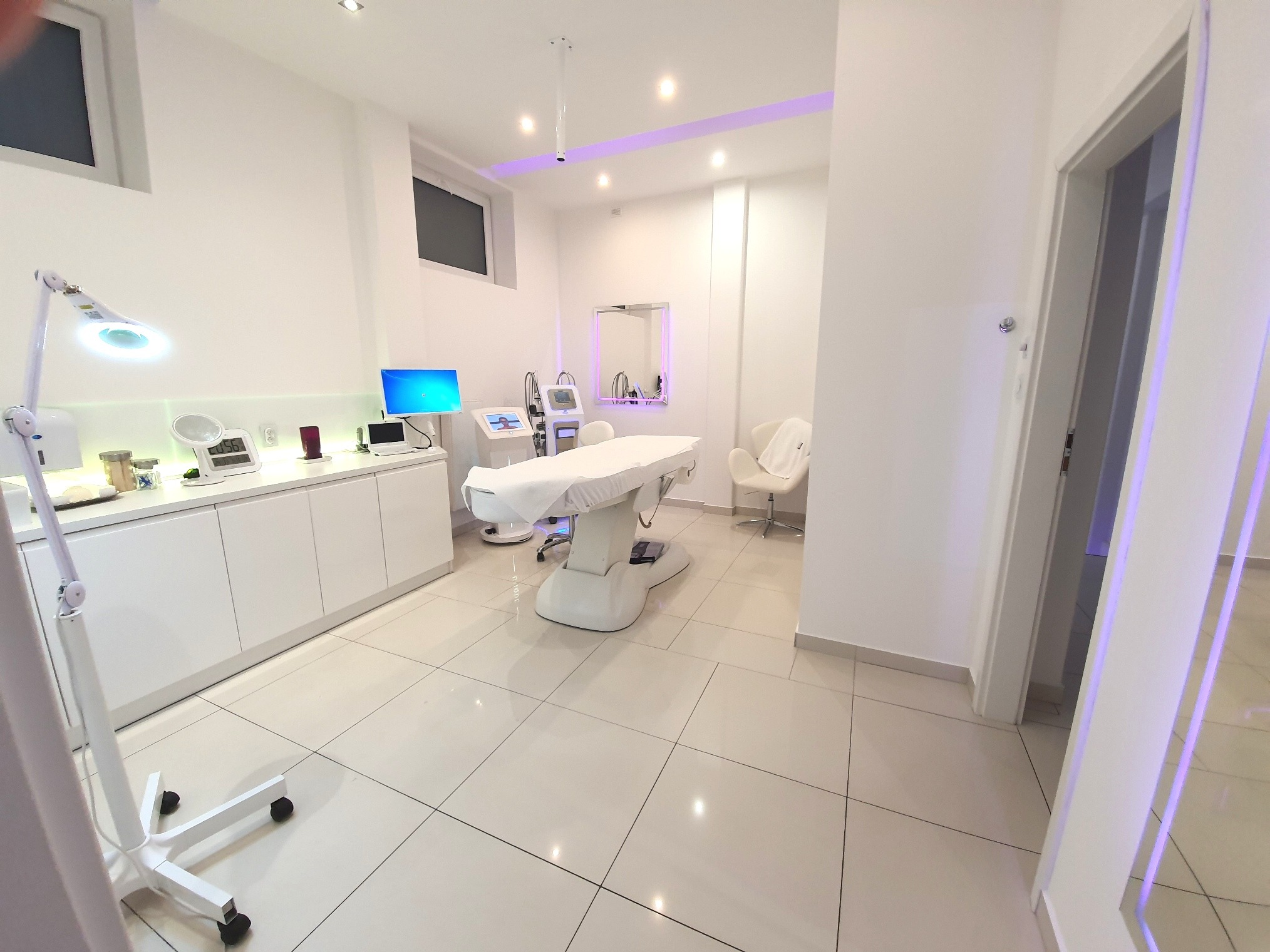 Jasne, przestronne wnętrze gabinetu z białymi meblami, sprzętem kosmetycznym i fotelem, oświetlone lampami sufitowymi i lampą podłogową z lupą.