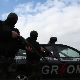 Dwóch uzbrojonych mężczyzn w kominiarkach, trzymających strzelby, stoi przed oznakowanym pojazdem z napisem 'GROM' i hasłem 'UZBROJONA FORMACJA OCHRONY'.