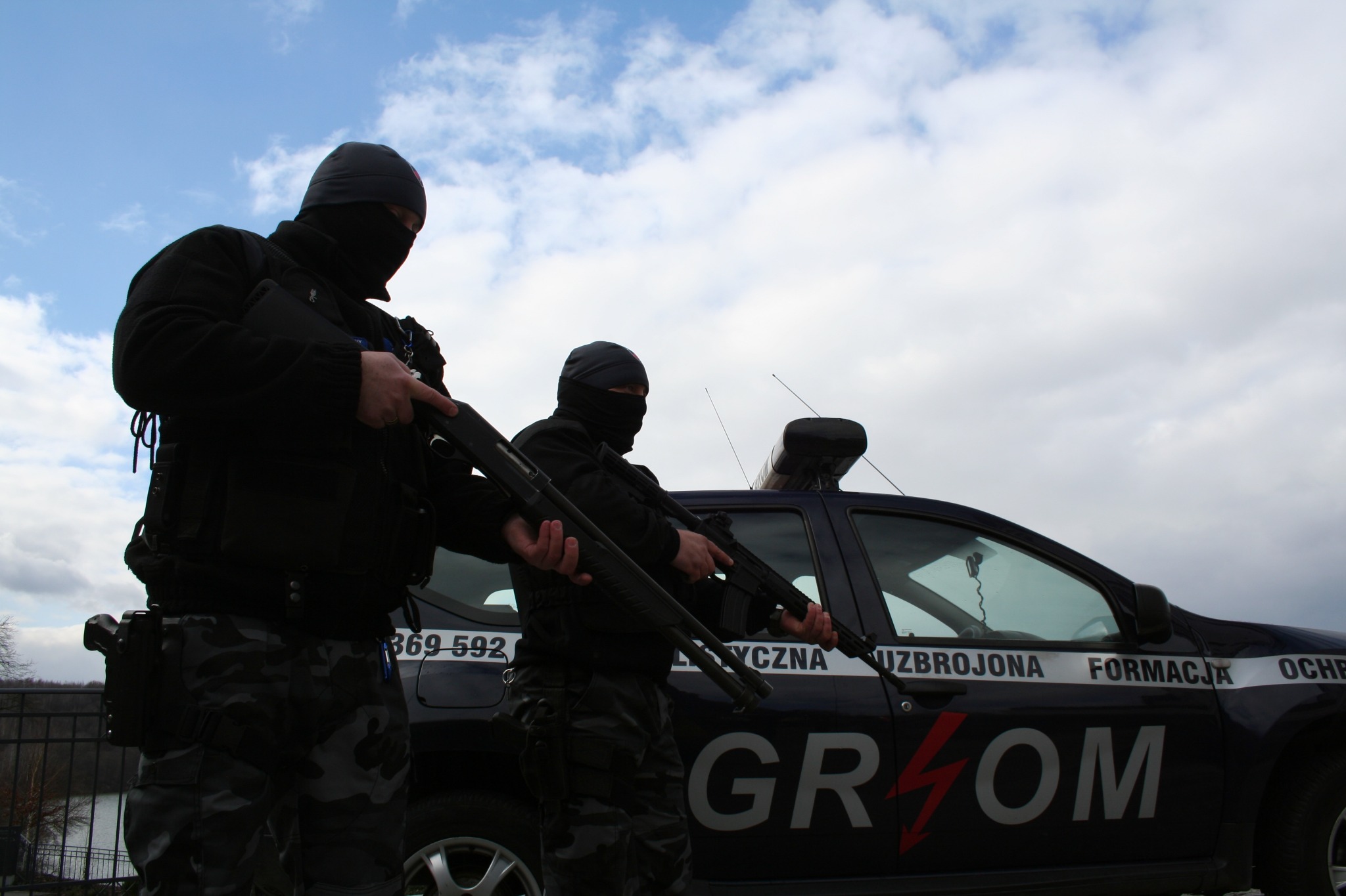 Dwóch uzbrojonych mężczyzn w kominiarkach, trzymających strzelby, stoi przed oznakowanym pojazdem z napisem 'GROM' i hasłem 'UZBROJONA FORMACJA OCHRONY'.
